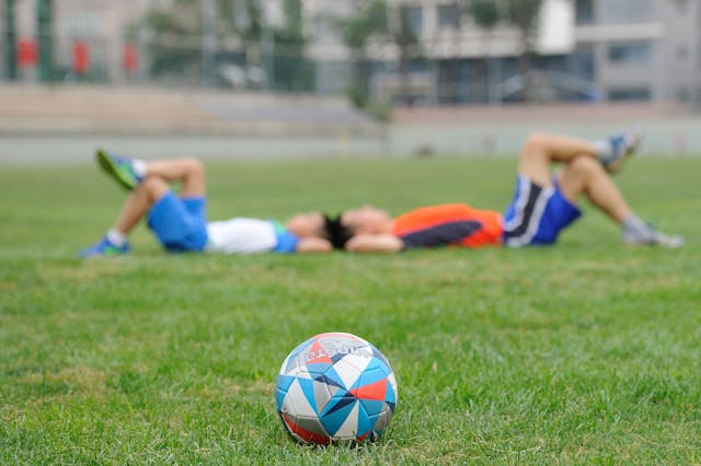 Balón de fútbol en primer plano con dos jóvenes descansando en el césped tras un partido en un campamento de verano.