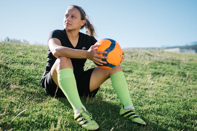 Jugadora de fútbol sentada sobre el césped con un balón en las manos y mirada decidida