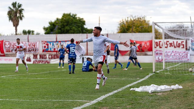 Futbolista celebrando un gol con euforia en un partido local, frente a las gradas y banderas de su equipo.